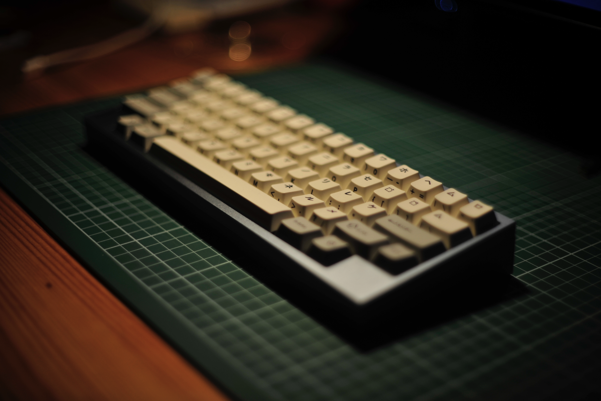 photograph of its new keyboard from the side, its a hhkb-layout mechanical board in a metal housing. the board has japanese (katakana) keycaps. the board sits on a cutting mat on a wood desk. only part of it is in focus.
